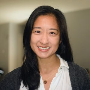 Portrait of a smiling woman with long black hair, wearing a white blouse and gray cardigan indoors.