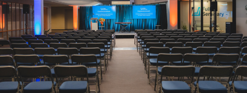 Conference setup in a bright atrium with rows of blue-cushioned chairs facing a stage with blue banners and a podium.